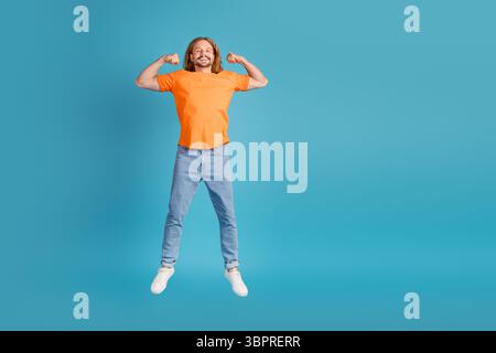 Jeune homme en t-shirt orange fléchissant ses bras en toute confiance sur un fond bleu vif Banque D'Images