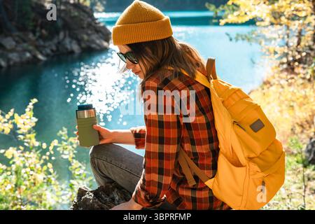 Jeune femme portant un bonnet jaune et chemise à carreaux est assis sur un rocher au bord d'un lac serein, tenant un thermos vert, entouré par le feuillage d'automne vibrant et Banque D'Images