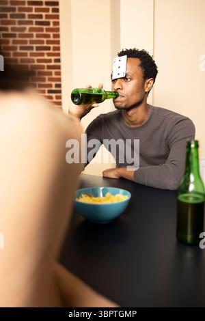 Jeune homme noir sirotant sa boisson pendant le voyage d'intérieur amusant avec des amis. Gars afro-américain assis à table avec carte à jouer sur le front, boire de la bière et profiter de moments de liaison avec les copains. Banque D'Images