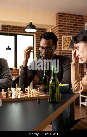 Homme afro-américain assis avec des amis, jouant aux échecs et grignotant dans la salle de mur de briques. Individu masculin noir pensif regardant l'échiquier, planifiant le prochain mouvement au cours d'un jeu de société amical intense. Banque D'Images