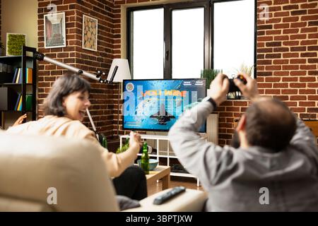 Homme blanc et femme se détendent ensemble, célébrant la victoire partagée du jeu vidéo. Jeunes adultes caucsasiens avec les mains levées, acclamant et dégustant des boissons sur le canapé tandis que la télévision fait clignoter le message gagnant. Banque D'Images