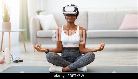 Femme afro-américaine portant des lunettes VR méditant faire du yoga en intérieur Banque D'Images