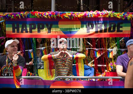 Une scène vibrante de défilé Pride avec un homme dans une chemise rayée arc-en-ciel soufflant un sifflet, donnant une forme de coeur avec ses mains. Une bannière « Happy Pride » Banque D'Images