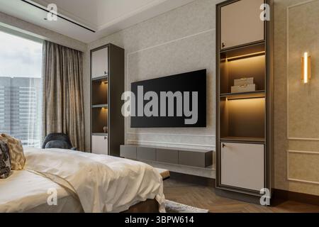 Chambre principale luxueuse et moderne avec un grand lit face à un mur de divertissement personnalisé. Comprend des étagères intégrées avec éclairage et une grande fenêtre. Banque D'Images