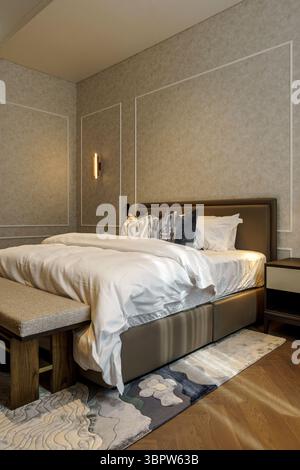 Une chambre sereine et luxueuse avec un lit soigneusement fait avec une couette blanche, un banc rembourré et d'élégants murs texturés avec moulures. Banque D'Images
