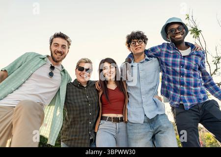 Un groupe joyeux et diversifié de jeunes amis adultes avec leurs bras l'un autour de l'autre, regardant directement la caméra et souriant ensemble à l'extérieur fr Banque D'Images