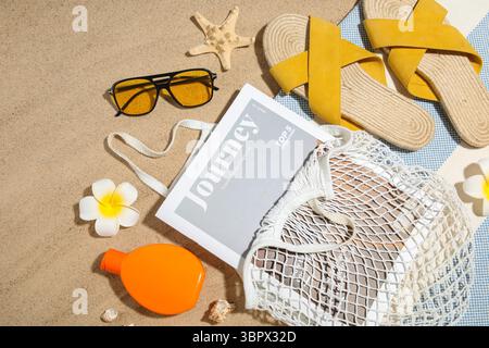 Sac à ficelle avec bouteille de crème solaire, magazine et accessoires de plage sur sable comme fond, gros plan Banque D'Images