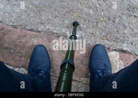 Un homme portant des chaussures bleues tient une canne de marche. Le bâton est vert et mesure environ la longueur de son bras. L'homme est debout sur un trottoir, et le Banque D'Images