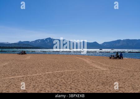 Des gens assis à des tables de pique-nique sur une plage de sable contemplant une eau bleue étincelante et des montagnes enneigées au lac Tahoe, en Californie Banque D'Images