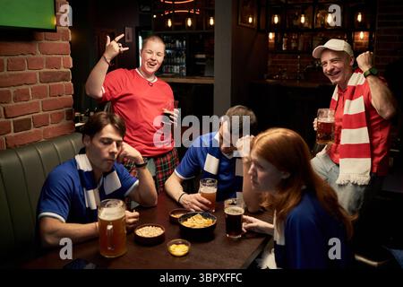 Groupe de fans de football multiethniques jeunes et d'âge moyen réagissant au match dans le bar, trois hommes caucasiens et une femme en maillots bleus regardant déçus, deux fans en rouge célébrant Banque D'Images
