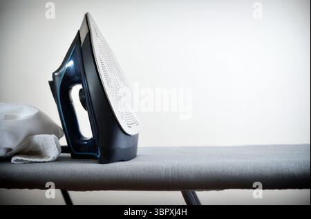 Concept de repassage de vêtements avec fer reposant verticalement sur une planche à repasser grise avec un vêtement blanc et un fond isolé léger. Vue de face. Banque D'Images