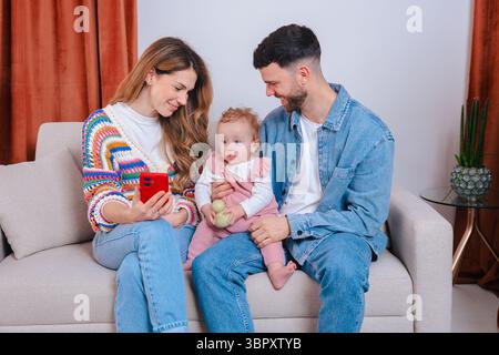 Les parents montrent le téléphone à bébé fille dans des combinaisons roses tout en se relaxant ensemble sur le canapé Banque D'Images