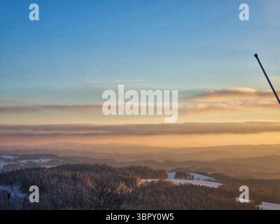 Le coucher de soleil hivernal vibrant peint le ciel nuageux orange et rose sur un paysage de montagne enneigé avec un pylône de remontée mécanique isolé dans une station de ski tchèque tranquille. Banque D'Images
