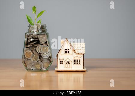 Pièces de monnaie dans un bocal en verre et petit modèle de maison sur une table, symbolisant l'épargne pour l'investissement de la maison ou de la propriété. Banque D'Images