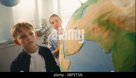 Classe STEM d'énergie renouvelable : garçon de l'école primaire attacher le modèle miniature de nuage au globe. Enseignante expliquant la science et les sources d'énergie à l'élève intelligent dans la salle de classe moderne. Gros plan. Banque D'Images