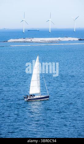 Yacht à voile dans les eaux bleues de Copenhague avec des éoliennes en arrière-plan par une journée ensoleillée, symbolisant l'énergie propre et le style de vie maritime. Banque D'Images