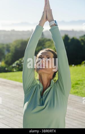 Femme mature étirant les bras au-dessus de la tête dans le haut athlétique sur la plate-forme en bois portant tracker de fitness. Bien-être, plein air, exercice, nature, vitalité, rela Banque D'Images