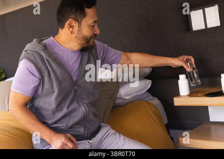 Homme asiatique assis dans la chambre à coucher atteignant des bouteilles de pilules et smartphone sur la table en bois, espace de copie. Décontracté, détente, moderne, style de vie, confort, wel Banque D'Images