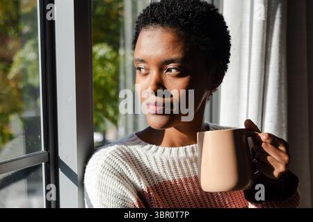 Femme afro-américaine tenant une tasse en céramique à la maison près de la fenêtre regardant le feuillage vert. Cosy, sérénité, détente, lumière naturelle, contemplatif, la vie Banque D'Images