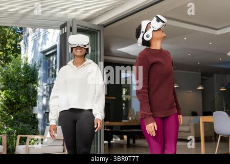 Diverses amies féminines debout côte à côte au seuil de la maison moderne portant des casques VR blancs. Technologie, innovation, casual, moderne, loisirs, o Banque D'Images