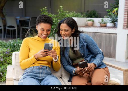 Diverses amies féminines vérifiant les écrans de smartphone tout en étant assis sur un canapé extérieur avec des plantes en pot. Détente, compagnie, Outdoorliving, lushpla Banque D'Images