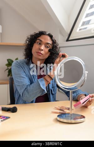 Personne non binaire appliquant le maquillage avec brosse au bureau loft près du miroir rond par puits de lumière, espace de copie. Créatif, maison, esthétique, style de vie, minimalisme, Banque D'Images