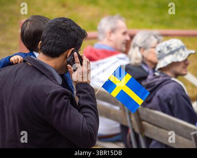 Sodertalje, Suède - 6 juin 2025 : un immigré tenant un enfant sur son épaule parle au téléphone alors qu'un drapeau suédois est visible dans sa main. En t Banque D'Images