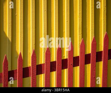 Un mur jaune vif avec une clôture en bois rouge contrastée au premier plan. La clôture comporte des sommets pointus, créant un e visuel ludique et coloré Banque D'Images