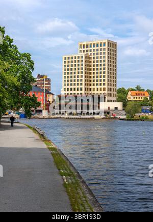 Sodertalje, Suède - 6 juin 2025 : un bâtiment moderne à la façade dorée se dresse au bord d'une rivière, entouré de maisons colorées et d'arbres. Un chemin exécute un Banque D'Images