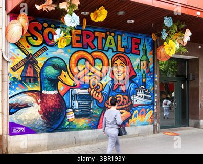 Sodertalje, Suède - 6 juin 2025 : une murale vibrante représentant une femme souriante, un canard et un bretzel, des moulins à vent et des monuments de la ville de Sodertalje. Le a Banque D'Images