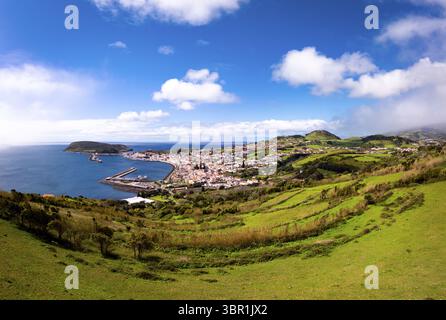 Vue aérienne de la ville nichée entre la mer bleue profonde et les collines verdoyantes, avec le port et Ponta da Espalamaca visible sous un ciel lumineux, Horta, Açores, Portugal. Banque D'Images