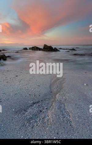 Vue sur la plage sereine où le ciel doux et pastel rencontre les rochers escarpés et les vagues douces, créant une scène côtière tranquille, Hermanus, Western Cape, Afrique du Sud. Banque D'Images