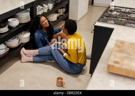 Femmes indiennes et afro-américaines assises sur le sol de la cuisine tenant smartphone près de la tasse à café. Amis, confortable, moderne, style de vie, domestique, occasionnel, guerre Banque D'Images