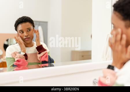 Femme afro-américaine touchant le visage examinant la peau dans le miroir à la vanité de salle de bain portant smartwatch. Beauté, soins de la peau, bien-être, autosoins, intérieur, m Banque D'Images