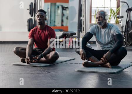Père et fils afro-américains méditant sur des tapis dans un centre de fitness portant des écouteurs. Bien-être, pleine conscience, force, connexion, vitalité, harmon Banque D'Images