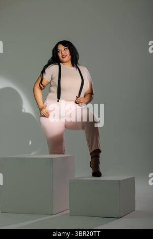Modèle féminin posant avec la jambe pliée sur le bloc blanc dans un studio minimaliste portant des bretelles noires. Mode, beauté, élégance, portrait, studio, visuel, m Banque D'Images