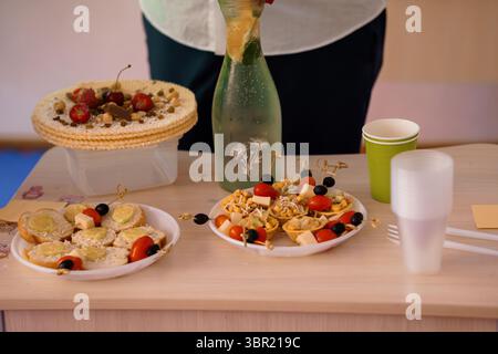 Tartinade colorée avec tarte aux fruits, brochettes de fromage et de tomates, en-cas à base de pain, boissons en bouteille et gobelets en plastique. nature morte, pique-nique intérieur Banque D'Images