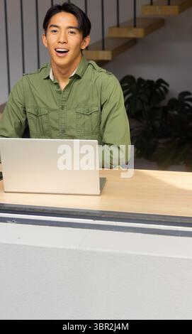 Homme asiatique portant chemise verte gestuelle à l'ordinateur portable sur le bureau dans le salon avec escalier flottant Banque D'Images