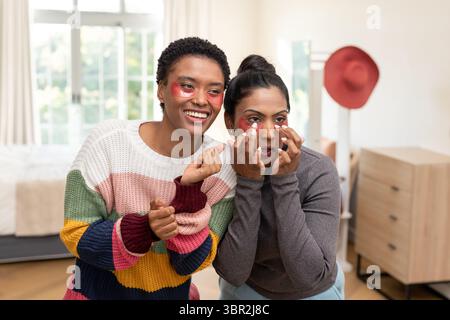 Diverses amies féminines appliquant des patchs rouges sous les yeux dans la chambre près du lit, porte-chapeau avec chapeau rouge Banque D'Images