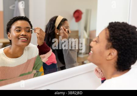 Diverses amies féminines appliquant un pinceau eyeliner et une baguette de mascara à la vanité devant le miroir Banque D'Images