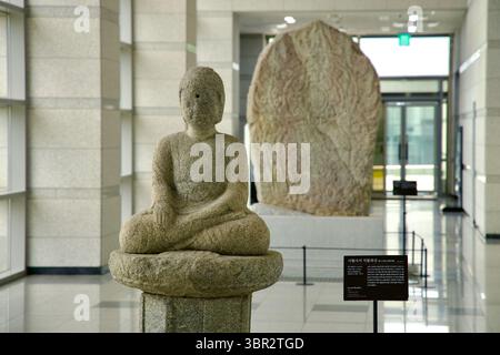 Gongju, Corée du Sud - 13 juin 2025 : une statue de Bouddha en pierre altérée se trouve à jambes croisées près d'une stèle sculptée dans le hall lumineux du musée national de Gongju Banque D'Images