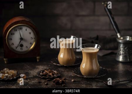 Thé ou café avec du lait et des épices en verre Armudu avec des grains de café et du sucre sur fond sombre Banque D'Images