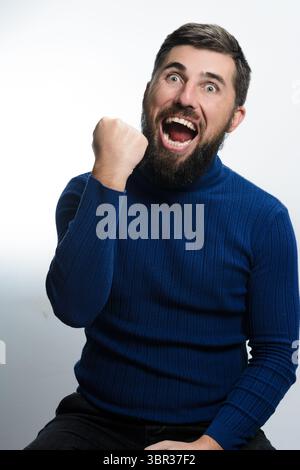 Un homme barbu dans un pull bleu et un pantalon sombre est assis sur un fond blanc, exprimant la joie avec un poing levé et la bouche ouverte. Banque D'Images