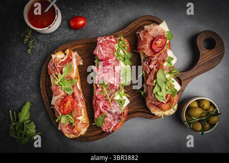 Assortiment de délicieux sandwichs baguette remplis de jambon ou de salami finement tranché et de laitue verte fraîche ou roquette pour le petit déjeuner disposés sur dos foncé Banque D'Images