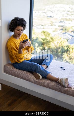 Femme afro-américaine assise sur le siège de la fenêtre à l'aide d'un smartphone avec des lunettes à côté, espace de copie Banque D'Images
