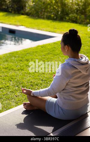 Femme asiatique méditant sur le tapis de yoga au bord de la piscine dans le jardin de l'arrière-cour portant smartwatch, espace de copie Banque D'Images
