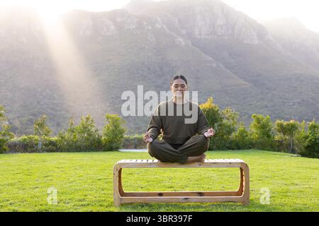Femme asiatique assise aux jambes croisées méditant sur un banc en bois dans le jardin surplombant la crête de la montagne Banque D'Images