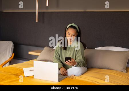 Femme asiatique assise sur un couvre-lit dans la chambre, tapant smartphone avec ordinateur portable et ordinateur portable à côté d'elle Banque D'Images