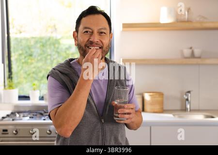 Homme asiatique mature debout dans la cuisine près du poêle plaçant la pilule dans la bouche et tenant le verre d'eau Banque D'Images