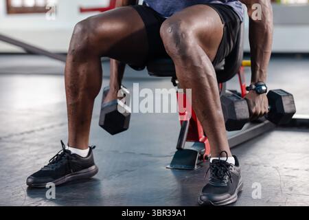 Homme afro-américain assis sur le banc rouge dans le gymnase portant une montre de fitness saisissant des haltères hexagonaux Banque D'Images